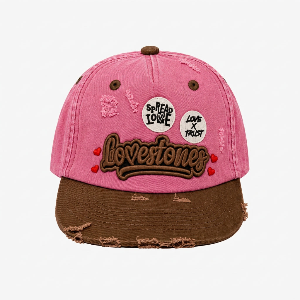 A pink and brown distressed baseball cap featuring 'Lovestones' embroidered on the front, two circular 'Spread Love' and 'Love x Trust' patches, and frayed details on the brim.